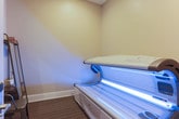 Located at 299 Harris Ln in Gallatin, Tennessee, this single-floor tanning salon offers a clean, comfortable, and professional environment designed for a relaxing tanning experience. The facility features a dedicated tanning room outfitted with a Luxura tanning bed, known for its high-quality UV light panels that emit a striking blue glow during operation. The room is finished with neutral, warm-toned walls and textured carpet flooring, creating a calm and private atmosphere for clients. A metal chair and a shelving rack provide convenient spots for personal belongings, and wall-mounted controls allow for easy session customization. The tanning room connects to a corridor finished with rich hardwood flooring and soft, neutral wall tones, lending a polished and welcoming feel to the overall space. Decorative framed artwork in the hallway adds a tasteful aesthetic touch. The layout is thoughtfully organized to ensure client privacy and comfort, with clear signage and accessible amenities throughout. The overall design strikes a balance between functional simplicity and a tidy, professional ambiance well-suited for a personal care or wellness business.