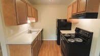 This single-floor residence presents a practical and straightforward layout designed for comfortable everyday living. The kitchen follows a galley-style configuration, with warm honey-toned wood cabinetry lining both sides of the corridor. Upper and lower cabinets provide generous storage space, while light-toned countertops offer a clean, functional workspace. A stainless steel sink sits along one wall, accompanied by a dishwasher, while the opposite side features a full suite of black appliances, including an electric coil range with an overhead exhaust hood and a full-sized refrigerator. Hardwood-style flooring runs throughout the kitchen, adding warmth to the space. The living area transitions to wall-to-wall carpeting in a neutral beige tone, contributing to a quieter, softer atmosphere underfoot. A large picture window along one wall is a standout feature, framing an outdoor view and allowing natural light to fill the room generously. A white paneled entry door with hardware provides a clean, classic entryway. The overall interior palette is neutral and understated, with light yellow-beige walls throughout, creating a cohesive and versatile backdrop suitable for a variety of furnishing styles.