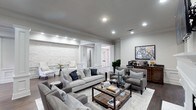 This single-story living space exudes a polished, contemporary elegance defined by thoughtful architectural detailing and a cohesive neutral palette. The main living area is generously proportioned, featuring crisp crown molding that frames the smooth grey ceiling, which is equipped with recessed lighting to provide layered, ambient illumination throughout the space.

A striking textured white stone accent wall serves as a focal point, adding depth and visual contrast to the otherwise refined interior. Decorative white columns and detailed millwork frame the entryway passage, lending a classic architectural character that complements the modern furnishings. Dark hardwood flooring runs throughout, grounding the space and contrasting beautifully with the light area rug beneath the seating arrangement.

The living area is thoughtfully arranged with a plush grey sofa, coordinating armchairs, and a rustic wood-and-glass coffee table, creating an inviting and functional gathering space. A dark wood console along one wall provides additional storage and display opportunities. The overall ambiance is one of understated luxury — bright, well-appointed, and carefully curated for both comfort and style.