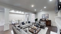This single-story living space exudes a polished, contemporary elegance defined by thoughtful architectural detailing and a cohesive neutral palette. The main living area is generously proportioned, featuring crisp crown molding that frames the smooth grey ceiling, which is equipped with recessed lighting to provide layered, ambient illumination throughout the space.

A striking textured white stone accent wall serves as a focal point, adding depth and visual contrast to the otherwise refined interior. Decorative white columns and detailed millwork frame the entryway passage, lending a classic architectural character that complements the modern furnishings. Dark hardwood flooring runs throughout, grounding the space and contrasting beautifully with the light area rug beneath the seating arrangement.

The living area is thoughtfully arranged with a plush grey sofa, coordinating armchairs, and a rustic wood-and-glass coffee table, creating an inviting and functional gathering space. A dark wood console along one wall provides additional storage and display opportunities. The overall ambiance is one of understated luxury — bright, well-appointed, and carefully curated for both comfort and style.
