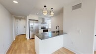 Located in the heart of Philadelphia's prestigious Rittenhouse Square neighborhood, this thoughtfully designed unit offers a clean, modern aesthetic throughout. Light hardwood flooring flows seamlessly across the living areas, complemented by crisp white walls and recessed lighting that create a bright, airy atmosphere.

The open-concept kitchen is a standout feature, centered around a sleek peninsula with a dark quartz countertop and an undermount sink. White cabinetry lines the walls, paired with stainless steel appliances including a French door refrigerator, electric range with a wall-mounted hood, and dishwasher. Two stylish pendant lights with black metal frames hang above the peninsula, adding a contemporary touch. A window in the kitchen brings in natural light and city views.

The spacious living room benefits from multiple large windows that frame expansive views of the Philadelphia skyline, bathing the space in abundant natural light throughout the day.

The full bathroom is finished with light tile flooring, a glass-enclosed marble-tiled shower, a floating white vanity with an undermount sink, and a window for natural ventilation and light. Matte black hardware accents throughout the unit add a refined, modern detail. A connecting hallway provides practical flow between all areas of the space.