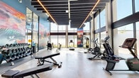 Located at 2121 Wood St in Oakland, CA, this contemporary fitness center offers a thoughtfully designed exercise space that combines industrial aesthetics with modern functionality. The room features soaring double-height ceilings adorned with a striking slatted black ceiling system and linear LED lighting strips that cast a warm, energizing glow throughout the space. Floor-to-ceiling windows wrap the perimeter, flooding the interior with abundant natural light and providing open views of the surrounding urban environment. The polished concrete flooring adds a sleek, durable finish that complements the overall industrial-modern design. Bold, colorful murals with Oakland-inspired graffiti art animate the accent walls, lending the space a vibrant and locally rooted character. The exercise room is well-equipped with a comprehensive selection of fitness machines, including treadmills, ellipticals, and stationary bikes for cardio training, alongside a cable machine, adjustable weight benches, and multiple dumbbell racks for strength training. The open floor plan allows for generous circulation between equipment zones, supporting a comfortable and efficient workout experience. A branded entry mat and clearly marked exit routes reflect the space's attention to safety and detail. Overall, the fitness center delivers a dynamic and inviting environment for a full range of exercise activities.