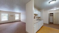 Located at 210 Washington Ave in Bridgeport, Connecticut, this single-floor unit offers a functional and straightforward layout designed for comfortable everyday living. The galley-style kitchen is well-appointed with white cabinetry lining both walls, ample counter space, a built-in dishwasher, electric range, refrigerator, and a sink beneath overhead cabinets — all finished with light hardwood-style flooring that adds warmth to the space. A pass-through window connects the kitchen to the living area, creating an open, communicative feel between the two spaces. The spacious living room features neutral carpet, textured ceilings, and baseboard heating, with windows that invite natural light and offer views of the surrounding greenery. The bedroom is generously proportioned, also finished with carpet and baseboard heating, and includes a large window dressed with blinds, framing outdoor foliage. The bathroom includes a full tub-shower combination with a clean white surround, a wood-toned vanity with a white countertop, a wall mirror, and hardwood-style flooring. Throughout the unit, the neutral color palette and clean finishes create a bright, move-in-ready atmosphere suited for a range of residents.