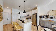 Located at 4027 Watercraft Ferry Ave in Wilmington, NC, this well-designed single-floor residence offers a cohesive blend of modern finishes and functional living spaces throughout. The kitchen serves as a central highlight, showcasing crisp white shaker cabinetry, granite countertops, a peninsula island with an undermount sink and seating for casual dining, and a full suite of stainless steel appliances including a range, microwave, and refrigerator. Stylish matte black pendant lights hang above the island, adding a contemporary contrast to the light, airy palette. Light wood-style flooring runs throughout the main living areas, enhancing the open and connected feel of the floor plan. The living room offers a comfortable, well-lit space with natural light filtering through slatted blinds, creating a bright and inviting atmosphere. The primary bedroom is generously sized, featuring a ceiling fan, walk-in closet, and direct access to an en-suite bathroom equipped with a dual-sink granite vanity, white cabinetry, and a large framed mirror with overhead lighting. An additional bedroom also benefits from natural light, a ceiling fan, and ample space. The overall design balances clean, modern aesthetics with warm, welcoming details throughout.