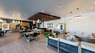 This contemporary community lounge is a thoughtfully designed amenity space that seamlessly blends form and function across a single, open floor. The interior is anchored by warm wood-look flooring that transitions into large-format slate-toned tile, clearly delineating the various activity zones within the space.

The main lounge area features a curated mix of modern furnishings, including rope-woven lounge chairs and plush sofas arranged beneath a dramatic slatted wood ceiling canopy. A standout feature is the bold geometric mural adorning one wall, rendered in deep teal, navy, gold, and taupe tones, which adds a striking artistic focal point to a dedicated lounge alcove furnished with low-profile seating.

The full kitchenette is well-appointed with an extended run of light ash wood cabinetry, a built-in beverage refrigerator, a professional-grade stainless steel refrigerator, a matte black utility sink, and a cooktop — all accented by a large-scale abstract painting mounted above. A bar-height counter with woven barstools provides casual seating directly adjacent.

A separate dining and social zone features a high-top communal table beneath a sculptural multi-globe chandelier. Floor-to-ceiling glass sliding doors flood this area with natural light and open directly to an outdoor patio, offering views of the surrounding community. Throughout, bold artwork, recessed lighting, and layered textures create a polished, gallery-like ambiance.