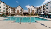 Located on Lime Quarry Road in Madison, Alabama, this outdoor amenity space is the central gathering hub of a modern residential community. The centerpiece is a generously sized resort-style swimming pool with crystal-clear water, framed by a wide concrete pool deck furnished with contemporary wave-shaped in-pool loungers and traditional chaise lounges positioned beneath market umbrellas. A covered pavilion anchors the poolside area, providing a shaded retreat equipped with seating and a relaxed outdoor living atmosphere.

Surrounding the pool, multiple distinct outdoor zones offer varied social settings. A landscaped courtyard features a built-in fire pit surrounded by stylish seating arrangements, accented by ornamental plantings, mulched garden beds, and string lighting overhead. Additional patio areas provide umbrella-shaded dining tables and curved lounge seating, encouraging casual outdoor gatherings throughout the day.

The community buildings, constructed with a combination of board-and-batten siding, brick masonry, and pitched rooflines, rise several stories and enclose the courtyard on multiple sides, creating a sense of enclosure and community. Mature plantings, decorative planters, and iron perimeter fencing contribute to the polished, resort-inspired ambiance of this outdoor space.