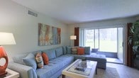 This single-floor space offers a welcoming and well-appointed interior designed for comfortable living. The living area serves as the centerpiece of the home, showcasing large sliding glass doors that flood the room with natural light and provide direct views of a verdant outdoor landscape beyond. The light-toned walls and textured ceiling complement the airy, open atmosphere throughout.

The living room is generously proportioned, accommodating a large tufted sectional sofa in a soothing blue-gray tone, accented with vibrant orange and multicolored throw pillows that add a pop of color and warmth. A modern glass-top coffee table anchors the seating arrangement, while a sleek floor lamp provides additional ambient lighting. A bold, colorful abstract artwork displayed on the wall adds a focal artistic element to the space.

The overall ambiance is bright, fresh, and inviting, with a cohesive color palette that balances cool and warm tones throughout. The layout is thoughtfully arranged to maximize both comfort and functionality, making the living space ideal for relaxation and everyday use. The seamless connection to the outdoors through the expansive glass doors enhances the sense of space and brings the natural surroundings into the interior experience.