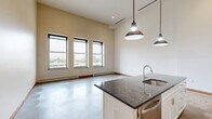 Located in downtown Flint, Michigan, this two-story loft unit at 200 E First St offers an open, industrial-inspired layout spread across two well-defined levels. The main floor is generously proportioned, showcasing polished concrete floors, soaring ceilings, and large black-framed windows that flood the space with natural light and frame views of the surrounding urban streetscape. The open-concept kitchen is a standout feature, equipped with white shaker cabinetry, stainless steel appliances including a French door refrigerator and range, a tile backsplash, and a substantial center island with a dark granite countertop, undermount sink, and stylish pendant lighting overhead. A modern cable-rail staircase leads to the upper loft level, which features carpeted flooring, recessed lighting, and a built-in closet with double doors — an ideal private retreat or flexible living space. The bathroom is thoughtfully finished with full-height tile walls, a marble-topped vanity with a barn-style sliding cabinet door, a wood-framed mirror, and warm tile flooring. Throughout the unit, warm wood trim accents provide contrast against the neutral palette, creating a cohesive and contemporary urban loft aesthetic.