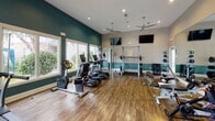 Located at 2402 N Ben Wilson St in Victoria, Texas, this well-appointed fitness center offers a comprehensive workout facility across a single open floor. The space is designed with a clean, modern aesthetic, featuring warm wood-style flooring, vaulted ceilings, and a two-tone color scheme of soft taupe and bold teal accents that lend a vibrant yet focused atmosphere.

Natural light pours in through multiple large windows, providing views of the surrounding outdoor greenery and creating a bright, energizing environment. Ceiling fans are strategically placed throughout to ensure comfortable air circulation during workouts.

The fitness floor is equipped with a variety of cardio machines, including treadmills, ellipticals, and stationary bikes. Strength training options include a power rack with barbells and weight plates, a cable resistance machine, a dumbbell rack, and a medicine ball station. Exercise balls are also available for functional training. Multiple wall-mounted flat-screen televisions are positioned for easy viewing from various areas of the room.

A full-length mirror spans one wall, maximizing the sense of space and supporting proper form during exercise. A dual-level water fountain is conveniently located near the exit for easy hydration access. The facility also displays a HealthHUB wellness information board, underscoring its commitment to resident health and well-being.