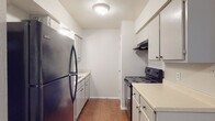 This single-story space is designed with a practical, functional layout evident throughout its rooms. The galley-style kitchen is efficiently organized, with white cabinetry lining both walls, light-toned countertops, and black appliances including a refrigerator and electric range with an overhead ventilation hood. Warm hardwood-style flooring runs the length of the kitchen, adding visual warmth to the crisp white cabinetry. A door at the far end of the kitchen provides additional access within the unit.

The bedrooms feature neutral carpet flooring and are finished with light-colored walls, creating a clean and understated atmosphere. Each room is equipped with a ceiling fan, contributing to air circulation and comfort. Natural light enters through windows fitted with blinds. One living area features wood-look tile flooring and a ceiling fan, providing a durable yet attractive surface.

The bathroom is well-appointed with a wide double-sink vanity topped with a light countertop, white cabinetry below for storage, a framed mirror, and a lighted vanity bar overhead. Wood-look flooring continues into this space, tying the areas together cohesively.

Overall, the space maintains a consistent, neutral palette with practical finishes throughout, creating a clean and move-in-ready environment.