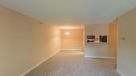 This single-floor space showcases an open-concept design that seamlessly connects the living area to the kitchen. The living space is finished with neutral-toned wall-to-wall carpeting, which adds warmth underfoot and complements the soft, light-colored walls throughout. The layout feels airy and spacious, offering flexible opportunities for furniture arrangement.

A peninsula-style kitchen counter is visible along the right side, featuring dark cabinetry and a set of appliances that includes a microwave, a stovetop range, and a refrigerator. The counter provides a natural division between the kitchen and the living area while maintaining the open feel of the space.

Overhead, a pendant light fixture hangs centrally in the dining or transitional area, providing focused ambient lighting and serving as a subtle visual anchor. Additional wall-mounted lighting is present in the living space. White baseboards run along the perimeter of the rooms, providing a clean finishing detail that contrasts neatly with the carpeted floors. Overall, the space presents a functional and understated aesthetic well-suited for comfortable everyday living.