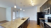 This single-floor space showcases a thoughtfully designed open-concept layout that seamlessly blends the kitchen and living areas. The kitchen is a standout feature, anchored by a generous island with light granite countertops and an undermount stainless steel sink. Dark espresso cabinetry with sleek hardware provides a striking contrast against the bright white walls, while warm hardwood-style flooring runs throughout, adding a sense of warmth and continuity to the space. Overhead, a combination of elegant pendant lights above the island and a modern track lighting fixture ensures ample illumination throughout. Stainless steel appliances, including a refrigerator and dishwasher, complete the kitchen's contemporary aesthetic. Natural light filters into the space through a glass-paneled door fitted with blinds, connecting the interior to the outside and contributing to the open, airy ambiance. The overall design is clean and modern, with neutral tones creating a versatile backdrop suited to a variety of interior styles. The open floor plan allows for flexible use of the living area adjacent to the kitchen, making the space feel expansive and well-connected.