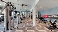 This fitness center is a spacious, single-floor facility designed to support a full range of workout routines. The layout is thoughtfully organized, dividing the space into dedicated zones for cardio and strength training. Along one side, a row of Cybex treadmills, elliptical trainers, and stationary bikes lines the windowed wall, allowing natural light to pour in while providing outdoor views during workouts. Ceiling fans are positioned throughout, contributing to a comfortable exercise environment. The strength training area features a robust selection of Hoist cable and resistance machines, a free weight station with a full dumbbell rack, and stability balls for functional training. Patterned carpet tile flooring covers the entire space, adding texture and durability underfoot. Recessed lighting complements the natural light, keeping the facility bright and well-illuminated. The walls are painted in a calming combination of soft blue and gray tones, creating a clean and motivating atmosphere. Wall-mounted flat-screen TVs provide entertainment for cardio sessions. Structural columns are integrated into the open floor plan without disrupting the flow of the space, making this fitness center a practical and inviting destination for daily exercise.