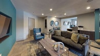 Located at 9240 Brukhaus in Raleigh, NC, this single-floor community space offers a sophisticated and welcoming environment defined by contemporary design and thoughtful layout. The grand entrance features floor-to-ceiling glass doors and windows with black frames, flooding the entryway with natural light and offering views of the surrounding neighborhood streetscape.

The main lounge area commands attention with dramatically soaring ceilings anchored by large stone-clad columns, pendant lantern chandeliers, and an abundance of natural light streaming through oversized windows dressed in patterned drapery. Large-format tile flooring flows throughout, lending a clean, cohesive aesthetic to the space. Seating areas are stylishly appointed with patterned accent chairs and glass-top tables, creating an inviting atmosphere for gathering.

A more intimate media lounge features a bold teal accent wall with a wall-mounted flat-screen TV, complemented by plush sofas and coordinating accent chairs. Adjacent to the main lounge, a sleek kitchenette offers dark cabinetry, quartz countertops, a built-in microwave, beverage refrigerator, and a striking textured tile backsplash. A bar-height work table positioned beside tall windows provides a bright spot for casual seating or workspace. The overall ambiance is modern, polished, and community-focused.