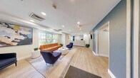 Located at 20 International Place in Tewksbury, MA, this thoughtfully designed amenity space offers a comprehensive set of facilities across a single floor. The welcoming reception lobby serves as the heart of the space, featuring light wood-look flooring, recessed lighting, and a sleek front desk finished in a soft stone-textured panel. Comfortable seating — including a tufted cognac leather sofa, navy accent chairs, and a round coffee table — creates an inviting lounge atmosphere, while large windows flood the area with abundant natural light and views of the surrounding greenery. A bold blue accent wall with vertical white stripe detailing leads into a well-lit corridor, adding a stylish, modern touch to the circulation path. The fitness center is generously proportioned, outfitted with multiple treadmills, a stationary bike, and additional cardio equipment, complemented by wall-mounted TVs, rubber flooring, and a window that brings in natural light. A dedicated office provides a private workspace, while a closet and bathroom round out the functional amenities. The overall design palette of cool blues, warm neutrals, and natural wood tones creates a cohesive, professional, and comfortable environment throughout.