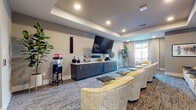 This dedicated screening and media lounge is thoughtfully designed for an immersive entertainment experience. The single-floor space centers around a large wall-mounted flat-screen television paired with a built-in soundbar, ensuring high-quality audio and visual performance. Rows of plush, yellow-patterned barrel chairs with attached side tables are arranged in a theater-style layout, providing comfortable, individual seating for multiple guests.

Anchoring the viewing wall is a run of sleek, dark navy cabinetry topped with a light granite countertop, offering generous storage alongside a built-in wine cooler. A classic popcorn machine stationed nearby adds a playful, cinematic touch to the ambiance. The room is finished with patterned, multi-tone carpeting that adds warmth and texture underfoot.

Acoustic wall panels in a deep slate blue line one side of the room, enhancing sound quality and contributing to the space's sophisticated aesthetic. A decorative glass-paneled door with a bold chevron inlay connects to an adjacent room, adding an artistic architectural detail. Natural light filters in through a curtained window, while recessed ceiling lighting and a tray ceiling detail create a polished, refined atmosphere throughout. Framed artwork on the walls completes the curated, upscale feel of this well-appointed media room.