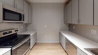 Located at 72 N Clinton Ave in the heart of downtown Rochester, NY, this single-floor unit offers a thoughtfully designed layout with modern finishes and a cohesive neutral color palette. Hardwood-style flooring runs throughout the space, complementing the soft gray walls and white trim that give every room a clean, contemporary feel.

The living area features soaring ceilings with a dramatic clerestory window that draws in natural light and adds architectural interest. An additional room benefits from two large windows with views of the surrounding urban streetscape, filling the space with daylight. Track lighting provides supplemental illumination where needed.

The galley-style kitchen is efficiently arranged with gray flat-front cabinetry lining both sides, generous white countertop surfaces, and stainless steel appliances including a range and built-in microwave, offering a streamlined cooking environment.

The bathroom is finished with a modern gray vanity, white countertop, a rectangular mirror with overhead lighting, and a walk-in shower featuring chrome fixtures and a clean white surround.

A dedicated closet with built-in shelving provides organized storage. The overall layout, as revealed by the floor plan, is elongated and functional, connecting each area with a logical flow suited to contemporary urban living.