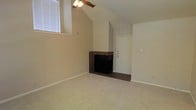 This single-story space features a welcoming living area defined by vaulted ceilings that lend a sense of openness and volume to the room. The dramatic ceiling height is complemented by a ceiling fan, which enhances air circulation and adds a practical touch to the space. Natural light filters in through a window positioned high on the wall, brightening the neutral-toned interior.

A brick fireplace serves as a notable focal point, adding character and warmth to the living area. Its traditional design contrasts with the clean, simple lines of the surrounding walls, creating visual interest. The fireplace surround features classic brickwork with a dark firebox interior.

The flooring throughout the living area consists of wall-to-wall carpeting in a light, neutral tone, contributing to a comfortable and understated aesthetic. The walls are finished in a consistent off-white palette, providing a versatile backdrop. A solid entry door is visible, offering a clear transition point to the exterior. Overall, the space presents a functional and comfortable layout with architectural details that add both practicality and charm.