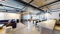 Located at 3 Executive Drive in Evesham, NJ, this single-floor amenity lounge delivers a compelling blend of industrial design and modern comfort. Exposed concrete ceilings with visible ductwork, steel beams, and track lighting establish a raw, loft-inspired aesthetic, while polished concrete floors unify the open-plan layout throughout.

A dramatic vertical metal slat partition serves as the space's defining architectural element, delineating zones without fully enclosing them, allowing natural light to filter freely across the floor. Large windows draped with sheer white curtains frame views of the surrounding wooded landscape, filling the interior with soft, diffused daylight.

The lounge area features plush sectional sofas, a low coffee table, and an area rug anchored beneath a wall-mounted display screen—ideal for relaxed gatherings. Adjacent to this, a full-size pool table takes center stage, with a dedicated cue rack and game accessories nearby, complemented by a framed Scrabble board display adding a playful touch. Arcade gaming units are tucked into a separate nook for additional entertainment.

A sleek bar and kitchen counter with barstool seating, fitted cabinetry, and a textured tile backsplash provides a functional refreshment station. The dining area accommodates multiple guests at round tables with contemporary upholstered chairs. Together, these zones create a versatile, social environment designed for both recreation and community engagement.