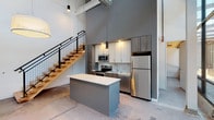This two-floor modern loft showcases an impressive industrial-chic aesthetic defined by soaring double-height ceilings, exposed ductwork, and polished concrete floors on the main level. The open-plan living area is anchored by a sleek kitchen equipped with stainless steel appliances, including a refrigerator, electric range, and microwave. White upper cabinetry contrasts with warm wood-toned lower cabinets, while a subway tile backsplash and quartz countertops add refined detail. A central island provides additional workspace and casual seating. Dramatic floor-to-ceiling steel-framed glass walls flood the main floor with natural light and offer views of the exterior courtyard. A striking open-tread staircase with natural wood steps and a black metal railing leads to the second floor, where light natural wood flooring extends throughout the bedroom spaces. The upper level continues the industrial-modern theme with large steel-framed windows that look out over the building's interior and beyond. Two bathrooms serve the unit — one on each floor — both fitted with wood-toned vanities, white quartz countertops, and clean, minimalist finishes. The upper bathroom includes a full tub-shower combination. The overall ambiance is airy, contemporary, and thoughtfully designed.