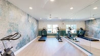Located at 2709 Pointe North Blvd in Albany, Georgia, this fitness center offers a well-appointed exercise space designed for a well-rounded workout experience. The room features light hardwood-style flooring that enhances the bright, clean aesthetic throughout. A striking artistic brick-pattern mural spans one full wall, rendered in teal and neutral tones, providing a visually engaging focal point that adds character to the space. Complementing this, a teal color-block accent band runs along the lower portion of the surrounding walls, tying the design together cohesively. Full-length mirrors line an entire wall, amplifying the sense of space and allowing users to monitor their form during workouts. Natural light filters in through multiple windows, keeping the environment open and inviting. Overhead, recessed lighting and a ceiling fan with light ensure the room remains well-lit and comfortably ventilated. The space is equipped with cardio machines including treadmills and ellipticals, a spin bike, a weight bench, dumbbell racks, medicine balls, and resistance training accessories, providing a comprehensive range of exercise options. A dark-framed glass door adds a modern touch to the entry. The overall ambiance is energetic yet polished, making this fitness center a functional and stylish amenity.