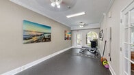 Located in Pensacola, FL, this dedicated fitness center offers a well-appointed exercise space designed to support a complete workout routine. The room features durable speckled rubber flooring throughout, providing both traction and comfort during exercise. Elegant crown molding and white baseboards lend a refined architectural character rarely found in a fitness facility.

Natural light flows in generously through a signature arched window and a glass-paned exit door at one end of the room, creating a bright, welcoming atmosphere. Ceiling fans and overhead fluorescent lighting ensure the space remains well-ventilated and evenly lit. A wall-mounted flat-screen television adds convenience for those looking to stay entertained during workouts.

The cardio zone is equipped with a treadmill, elliptical trainer, and stationary bike, while the strength training area features a dumbbell rack, adjustable weight bench, and kettlebells. Resistance cables are also available for varied training. A large framed mirror aids in form correction, and a sanitation dispenser supports cleanliness and hygiene.

Vibrant coastal-themed canvas artwork adorns the walls, contributing to an energizing yet relaxed ambiance. French doors connect to an adjacent space, and views of lush greenery through the windows reinforce the Florida setting. This fitness center is a functional and attractively designed space for residents seeking convenient on-site exercise amenities.