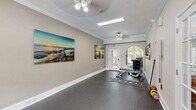 Located in Pensacola, FL, this dedicated fitness center offers a well-appointed exercise space designed to support a complete workout routine. The room features durable speckled rubber flooring throughout, providing both traction and comfort during exercise. Elegant crown molding and white baseboards lend a refined architectural character rarely found in a fitness facility.

Natural light flows in generously through a signature arched window and a glass-paned exit door at one end of the room, creating a bright, welcoming atmosphere. Ceiling fans and overhead fluorescent lighting ensure the space remains well-ventilated and evenly lit. A wall-mounted flat-screen television adds convenience for those looking to stay entertained during workouts.

The cardio zone is equipped with a treadmill, elliptical trainer, and stationary bike, while the strength training area features a dumbbell rack, adjustable weight bench, and kettlebells. Resistance cables are also available for varied training. A large framed mirror aids in form correction, and a sanitation dispenser supports cleanliness and hygiene.

Vibrant coastal-themed canvas artwork adorns the walls, contributing to an energizing yet relaxed ambiance. French doors connect to an adjacent space, and views of lush greenery through the windows reinforce the Florida setting. This fitness center is a functional and attractively designed space for residents seeking convenient on-site exercise amenities.