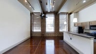 This single-floor loft space masterfully blends industrial character with modern functionality. The open layout is anchored by rich, polished concrete floors that gleam under the light, reflecting the warmth of the exposed brick walls that line the perimeter. Large, grid-paned windows dressed with horizontal blinds allow generous amounts of natural light to pour into the space, creating a bright and welcoming atmosphere throughout the day. The soaring ceiling adds a dramatic sense of volume and openness, enhanced further by exposed ductwork and structural beams that celebrate the space's industrial roots. A ceiling fan with a bold black finish adds both a utilitarian and stylistic touch overhead. Track lighting runs along the ceiling, offering flexible and directional illumination suited for a variety of uses. On one side, a built-in counter with shelving provides a practical workspace or serving area, accompanied by modern pendant lighting. The combination of raw materials — brick, concrete, and metal — alongside clean white walls creates a compelling contrast that gives this space a distinctive, loft-style character full of personality and potential.