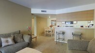This single-floor residence features an open-concept layout that seamlessly connects the living, dining, and kitchen areas into one cohesive space. The interior is finished in warm beige and cream tones, with carpeted flooring throughout the main living areas that contributes to a comfortable, understated ambiance.

The living area is furnished with upholstered seating and a small coffee table, providing a relaxed lounge space. A dedicated dining area accommodates a modest table with wooden chairs, positioned near framed wall art that adds a decorative focal point to the room. Natural and recessed lighting keeps the space well-lit and welcoming.

The kitchen is defined by a raised breakfast bar with metal barstool seating, creating a casual countertop dining option. Light wood cabinetry lines the kitchen walls, and a built-in microwave is visible above the cooking range, offering practical functionality. The bar counter creates a subtle division between the kitchen and the living areas while maintaining the open feel of the overall floor plan.

A hallway visible toward the rear of the space leads to additional rooms, suggesting private areas beyond the shared living zones. The layout is efficient and well-organized throughout.