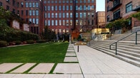 Located at 1296 W Sixth Street in Cleveland, Ohio, this outdoor courtyard space sits nestled within a striking complex of historic red brick industrial buildings in the heart of the Warehouse District. The courtyard masterfully blends preserved architectural heritage with thoughtful modern landscaping design.

The hardscape features expansive large-format stone pavers that create a clean, contemporary walkway and gathering area. Raised stone and concrete planters house mature trees that provide natural shade and greenery, softening the industrial character of the surrounding brick facades. Sleek black metal railings define level transitions and guide movement through the space, while broad stone steps connect varying elevations seamlessly.

A lush green lawn panel, bordered by stepping stone pathways, adds a natural, park-like quality to one section of the courtyard. Warm wood-toned pergola elements and built-in seating areas further enhance the outdoor experience, offering areas for rest and gathering. The multi-story brick buildings that enclose the courtyard feature large multi-pane industrial windows that flood the surrounding facades with character. The interplay of weathered brick, stone, wood, and mature plantings creates a welcoming urban oasis that honors the site's industrial past while providing a refined, functional outdoor environment.