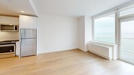 Located at 1 Ocean Dr in Brooklyn's Coney Island neighborhood, this modern one-bedroom, one-bathroom residence offers a thoughtfully designed layout across a single floor. Light-toned hardwood flooring flows throughout, complementing the clean white walls and contemporary finishes that define the space.

The open-plan kitchen and living area serves as the heart of the unit. The kitchen is outfitted with high-gloss white cabinetry extending floor to ceiling, a textured wave-pattern tile backsplash, stainless steel appliances including a gas range, microwave, dishwasher, and refrigerator, and a undermount sink set into a white quartz countertop. Large windows in the living area frame open water views, filling the space with natural light.

The bedroom is a bright, comfortable retreat featuring wide windows with sweeping ocean views, light hardwood flooring, and a closet. The unit includes a total of four closets, offering generous storage throughout. The bathroom makes a striking impression with floor-to-ceiling marble-style wall tile, a wood-accented vanity, and a white undermount sink. A dedicated laundry area and a private balcony round out the unit's practical amenities, making this a well-appointed coastal residence.