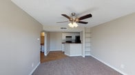 This single-story space showcases an open-concept design that seamlessly connects the living area to the kitchen. The living room is finished with plush carpet flooring and neutral beige walls, creating a warm and inviting atmosphere. A ceiling fan with a built-in light fixture is centrally mounted on the textured ceiling, providing both air circulation and ambient lighting.

A standout feature of the living area is the built-in shelving unit along one wall, offering practical display and storage space. The kitchen is accessible via the open layout and features white cabinetry, dark countertops, and black appliances, including a refrigerator and range. A peninsula-style counter serves as a natural divider between the kitchen and living space, ideal for casual dining or additional workspace.

The kitchen transitions to hardwood-style flooring, providing a clear visual distinction between spaces. An arched doorway leads to an additional room, visible in the background with its own flooring and lighting. The overall design balances functionality with a clean, neutral aesthetic suitable for a variety of interior styles.