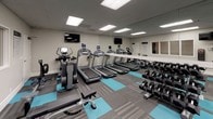 Located at 225 W 3rd Street in Long Beach, California, this fitness center offers a well-appointed, single-floor workout facility designed to support a full range of exercise routines. The space features a contemporary aesthetic, with a distinctive patterned carpet in grey and teal tones that adds visual energy to the room.

The cardio zone is equipped with multiple treadmills and elliptical machines, each outfitted with individual digital displays and conveniently positioned wall-mounted flat-screen televisions, allowing users to stay entertained during their workouts. The strength training area includes a cable machine with a functional training bench, a full rack of dumbbells ranging across various weights, and additional free weight accessories such as medicine balls and stability balls.

Floor-to-ceiling mirrors line the walls throughout the space, creating a sense of openness and allowing users to monitor their form. Recessed panel lighting in the drop ceiling provides bright, even illumination across the entire facility. A separate section of the space features warm hardwood flooring, suggesting a dedicated stretching or functional movement area. Spin bikes and other specialty equipment round out the offerings, making this a comprehensive fitness destination in the heart of Long Beach.