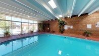 Located at 2200 Lester Dr NE in Albuquerque, New Mexico, this single-floor indoor pool facility offers a well-appointed aquatic environment designed for comfort and leisure. The centerpiece of the space is a generously sized rectangular swimming pool with clearly marked depth indicators, filled with vivid turquoise water. The pool deck is finished in a distinctive terracotta-red surface that wraps around the perimeter, providing ample space for lounging with scattered chairs and seating arrangements. Exposed white structural beams run across the ceiling, complemented by multiple skylights that allow natural light to filter in, creating a bright and open atmosphere. A full wall of floor-to-ceiling glass windows and sliding doors lines one side of the room, offering views of an outdoor patio area and the surrounding landscape, further enhancing the sense of openness. The opposite wall features classic exposed brick, adding warmth and texture to the interior. Lush potted plants and climbing greenery are distributed throughout the space, softening the architectural elements and contributing to a welcoming, resort-like ambiance. Safety equipment, including life rings, is mounted near the poolside windows. An adjacent glassed-in room is visible through the interior windows, suggesting additional amenity spaces connected to this facility.