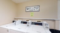 Located at 421 Iverson Way in Galloway, Ohio, this single-floor self-serve laundry facility offers a clean and practical environment designed for convenient, efficient use. The laundry room features a row of commercial-grade Speed Queen washers positioned along a neutral-toned wall, providing users with reliable, high-capacity machines suited for a variety of laundry needs. A wall-mounted shelf runs above the machines, offering a convenient surface for laundry supplies and personal items. The space is accented by a decorative "Wash and Dry Laundry Co." sign, lending a welcoming, classic aesthetic to the otherwise functional room. The walls are finished in a soft, light tone that contributes to a bright and tidy atmosphere. The facility is straightforward in its layout, making it easy to navigate and use. Informational notices are posted on the wall, keeping users informed of any relevant guidelines or instructions. Overall, this laundry facility is well-maintained and thoughtfully equipped, providing a dependable and accessible space for self-serve laundry needs in the Galloway community.