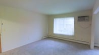 This single-floor space features a simply designed room with smooth, light-colored walls and wall-to-wall carpet in a neutral tone, providing a clean and versatile foundation. A large window fitted with vertical blinds serves as a central architectural feature, allowing an abundance of natural light to fill the room and creating a bright, open atmosphere. The window offers a view of the exterior surroundings, adding a connection to the outdoors. A baseboard heating unit runs along the base of the exterior wall beneath the window, ensuring efficient climate control throughout the space. The walls are finished in a soft, pale yellow-white tone that complements the light carpeting, contributing to the room's airy and understated ambiance. The overall design is straightforward and functional, with smooth ceilings and minimal ornamentation, making the space highly adaptable to a variety of uses and furnishing styles. The neutral palette and natural light make it a welcoming and practical environment.