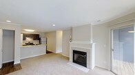 Located at 1100 Audubon Parc Dr in Cary, North Carolina, this single-floor residence offers a thoughtfully designed layout with a warm, neutral aesthetic throughout. The open-concept living area serves as the heart of the home, anchored by a classic white-mantled fireplace with white tile surround, adding both visual appeal and a cozy focal point to the space. Soft, wall-to-wall carpeting in a warm beige tone extends across the living area, contributing to a comfortable and inviting ambiance. The walls are finished in a light, neutral palette that complements the bright, airy feel of the space. Recessed lighting provides clean, even illumination throughout. The kitchen is seamlessly connected to the living area and features dark cabinetry, a light stone-finish countertop, stainless steel appliances including a refrigerator and range, and an under-cabinet light fixture that enhances functionality. A hallway visible from the living area suggests additional rooms beyond, offering a sense of depth and privacy within the floor plan. Overall, the space presents a clean, move-in-ready environment with practical finishes and a cohesive design sensibility.