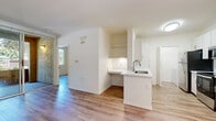Located at 9320-9370 Hillery Dr in San Diego's Mira Mesa community (92126), this single-floor unit offers a clean, modern aesthetic defined by light-toned wood-look flooring and a bright, neutral color palette throughout. The updated kitchen is a standout feature, showcasing white shaker-style cabinetry, stone-look countertops, a stainless steel refrigerator, dishwasher, and range, along with an in-unit laundry area conveniently accessible from the kitchen corridor. A peninsula with a sink and additional counter space connects the kitchen to the dining area, which benefits from generous natural light through a large window accented by a pendant chandelier. The living area flows comfortably from the kitchen, offering access to an enclosed patio or balcony through sliding glass doors, bringing in further natural light and outdoor views of lush greenery. The bedrooms are well-proportioned, each featuring wood-look flooring, neutral walls, and ample natural light from windows overlooking the surrounding landscape. Crown molding adds a refined touch to the living and bedroom spaces. The bathroom is outfitted with a tiled tub-shower combination, white vanity with a clean countertop, and matching wood-look flooring, maintaining the cohesive, contemporary design found throughout the unit.