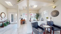This well-appointed single-floor space offers a warm and professional atmosphere, characterized by vaulted ceilings, skylights, and light-toned hardwood flooring that runs throughout. The entry foyer welcomes visitors through glass-paneled double doors, flanked by sidelight windows that flood the space with natural light. Exit signage and wall sconces provide both safety and ambient illumination, while the soaring ceiling height adds an open, airy quality from the moment of arrival.

Beyond the entry, the layout opens into a reception and waiting area, where a half-wall partition with a service counter separates staff and visitor zones. The waiting area is furnished with leather accent chairs and a side table, offering a comfortable and professional setting. Decorative wall art and sconce lighting contribute to the refined ambiance.

A dining or conference area sits adjacent, bathed in natural light from a wide bank of windows that offer views of an outdoor pool area. A round wooden table with spindle-back chairs anchors this space, making it ideal for meetings or informal gatherings. A prominent potted tree adds a natural, refreshing touch to the corner.

A secondary workstation area is also visible, featuring a wooden desk, small appliances, and additional window views to the exterior. Throughout the space, the neutral color palette, clean architectural lines, and thoughtful use of natural light create a cohesive, inviting environment suited for professional or community use.
