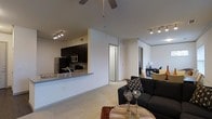 Located in Richardson, this single-floor unit showcases a thoughtfully designed open-concept layout that seamlessly connects the kitchen and living areas. The kitchen is appointed with dark cabinetry, stainless steel appliances including a refrigerator and microwave, a decorative tile backsplash, and a granite-topped breakfast bar that provides both additional prep space and casual seating. Track lighting illuminates the kitchen, while a ceiling fan anchors the living space, promoting airflow and comfort. The living area is generously proportioned, featuring plush carpeting underfoot and neutral-toned walls that create a clean, versatile backdrop. Soaring ceilings enhance the sense of openness throughout the space. A hallway leads to additional rooms, with a glimpse of a secondary space visible in the background, suggesting flexible use for a bedroom, office, or dining area. The overall ambiance is modern and inviting, with a consistent design language that ties each area together. The combination of practical finishes and contemporary styling makes this a highly functional and visually appealing space suited for everyday living.