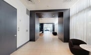 Located at 3635 NE 1st Ave in Miami's vibrant Design District, this contemporary amenity floor at Eve at the District offers a thoughtfully designed, single-level space spanning a generous footprint. The layout flows seamlessly from a striking entrance corridor clad in large-format dark tile panels, which frame an elegant passageway leading deeper into the space.

The main gathering area features a curated lounge with floor-to-ceiling built-in bookshelves set against bold teal-blue backing, complemented by a glass-top communal table surrounded by classic white Eames-style chairs atop warm hardwood flooring. Vibrant artwork adds a creative energy to this zone, making it ideal for both casual socializing and focused work.

A dedicated yoga studio is accessible via dramatic script-etched glass doors, offering a private, light-filled retreat. Adjacent to this, a bright corridor runs alongside expansive floor-to-ceiling windows that flood the space with natural light and provide views of an outdoor terrace featuring lush tropical plantings and Miami's skyline beyond.

The entrance is marked by the Eve at the District branded mat and sheer drapery, creating a polished, welcoming arrival experience. Recessed lighting throughout maintains a clean, modern ambiance, while the consistent use of light-toned tile flooring, dark accent panels, and crisp white walls unifies the entire space with a sophisticated, urban aesthetic.
