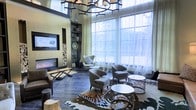 This elegantly designed lounge space makes an immediate impression with its dramatic double-height ceilings and expansive floor-to-ceiling windows dressed in sheer white curtains that bathe the interior in soft, natural light. An eye-catching wrought iron chandelier with an organic, branch-like form anchors the ceiling and serves as a bold architectural focal point.

The main seating area is thoughtfully arranged around a sleek, wall-mounted linear fireplace, which is flanked by a dark metal shelving unit displaying curated decorative accents. Rich, dark hardwood floors run throughout, grounding the space with warmth and contrast. A striking cowhide-patterned area rug in earth tones defines the central gathering zone, where an assortment of upholstered accent chairs in neutral grey tones invite relaxed conversation.

Complementing the seating arrangement are sculptural side tables with metallic bases and clean white surfaces, adding a modern, artistic touch. A large wall-mounted flat-screen display is integrated seamlessly into the design near the fireplace wall. The overall ambiance strikes a balance between rustic and contemporary, with natural textures, layered lighting, and a carefully curated palette of warm neutrals creating an inviting and refined atmosphere throughout.