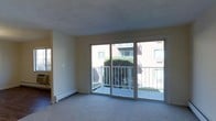 This single-floor residential space offers a clean, functional layout designed for comfortable everyday living. The living area is a standout feature, highlighted by expansive sliding glass doors that flood the room with natural light and provide direct access to a private balcony. The balcony is enclosed by a white metal railing and overlooks a landscaped courtyard with mature greenery, offering a pleasant outdoor retreat. The living space is finished with wall-to-wall carpet and neutral-toned walls, creating a calm and versatile backdrop suited to a variety of interior styles. A baseboard heating unit runs along the exterior wall, ensuring efficient climate control throughout the colder months. The open sightlines between rooms contribute to an airy, connected feel within the unit. Additional windows allow natural light to reach adjacent areas, further enhancing the brightness of the interior. Overall, the space presents a straightforward and practical residential layout, well-suited for those seeking a comfortable, light-filled home with convenient outdoor access.