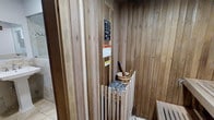 This sauna is constructed entirely with tongue-and-groove cedar wood paneling, covering the walls, ceiling, and benches to create a warm, cohesive aesthetic. The natural wood grain is prominently visible throughout, lending an authentic Scandinavian-style atmosphere to the space. At the center of the room sits a traditional rock sauna heater, encased in a slatted wooden safety guard to prevent accidental contact. A classic wooden bucket and ladle rest beside the heater, ready for water to be poured over the rocks to generate steam. Safety and usage instruction placards are affixed to the wall near the entrance, providing guidance to users. The flooring transitions from tile just outside the sauna door to wooden slat benches within, typical of a properly designed wet or dry sauna environment. The compact interior is designed to retain heat efficiently, with a tightly fitted door and well-insulated cedar walls. The overall ambiance is rustic yet functional, offering a traditional sauna experience suited for relaxation and therapeutic heat therapy.