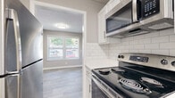 This single-floor residence offers a clean, modern aesthetic with a cohesive neutral color palette of soft grays and crisp whites throughout. The kitchen is a standout feature, outfitted with white shaker-style cabinetry, white subway tile backsplash, white quartz countertops, and a full suite of stainless steel appliances including a refrigerator, electric range, and over-the-range microwave. Light wood-look flooring runs through the kitchen and flows seamlessly into the open living and dining areas, which are brightened by natural light from multiple windows. The living space connects easily to a hallway leading to the private quarters of the unit. The bedrooms feature plush neutral carpeting, textured ceilings, and windows that bring in additional natural light. The bathroom is thoughtfully updated with a wide white vanity topped with a white quartz countertop, an undermount sink with a brushed nickel faucet, and a gold-framed mirror as a refined accent. A tub-shower combination with tile surround and wood-look tile flooring completes the bathroom. Throughout the space, white trim and baseboards provide clean contrast against the warm gray walls, contributing to a polished and move-in-ready atmosphere.