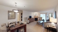 This single-floor residence offers a thoughtfully designed open-concept layout that seamlessly connects the dining and living areas, creating a spacious and comfortable environment. The dining area is anchored by a rich dark wood table set beneath an elegant pendant light fixture, with a framed nature-inspired artwork adorning the wall nearby. Neutral-toned carpeting flows throughout the space, contributing to a warm and inviting ambiance. The living area is generously appointed with comfortable seating, including wingback chairs arranged around a central coffee table, and features a corner fireplace that serves as a cozy focal point. A media console with a mounted television provides functional entertainment options. Natural light fills the room through a large sliding glass door, which opens to an outdoor area and offers pleasant exterior views. A built-in shelving area is visible near the entryway, adding practical storage to the layout. The overall design palette of soft whites, warm beiges, and wood tones creates a cohesive, serene atmosphere throughout the residence.