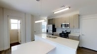 This contemporary two-floor residence is designed with clean lines and a modern aesthetic throughout. The kitchen serves as a central highlight, showcasing an expansive white countertop island that doubles as a prep and gathering space, paired with an undermount sink and modern pendant lighting overhead. Flat-panel cabinetry in a warm neutral tone provides ample storage, while a decorative tile backsplash adds texture and visual interest to the space. Stainless steel appliances — including a gas range, built-in microwave, and French door refrigerator with an ice dispenser — complete the professional-grade kitchen setup. Recessed and overhead lighting ensures the space is well-lit, while a window at the far end invites natural light into the room. The overall color palette of white, beige, and stainless steel creates a bright, cohesive, and functional environment. The two-floor layout suggests additional living, sleeping, or utility spaces beyond the kitchen, offering a well-rounded and versatile floor plan suited for everyday living.