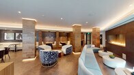 This thoughtfully designed single-floor amenity lounge combines contemporary aesthetics with warm, natural materials to create an inviting and versatile communal environment. The space is anchored by richly textured wood-clad columns that define distinct zones throughout the open floor plan, all unified by large-format stone tile flooring and a seamlessly lit tray ceiling with recessed and cove lighting.

At the heart of the space, a stylish lounge area features bold, patterned barrel chairs alongside sleek light-blue curved sofas, offering a striking contrast of patterns and tones. A wall-mounted linear gas fireplace set into a dark mosaic tile surround adds warmth and a dramatic focal point to one end of the space.

A generous communal bar counter runs along a window-lined wall, flooded with natural light and outfitted with bar-height seating. Potted greenery lines the counter, and large windows frame views of an adjacent outdoor area, creating a bright and airy atmosphere. Curated artwork adorns the walls throughout.

A dedicated dining nook features upholstered banquette seating, natural wood booth tables, and pendant lighting, providing a more intimate setting. Planters filled with lush snake plants and succulents are positioned near entryways, enhancing the biophilic design sensibility that runs throughout the space.