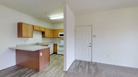 This single-floor unit offers a practical and well-organized layout designed for comfortable everyday living. The entryway greets you with light wood-look flooring and a security panel, providing a welcoming transition into the main living areas. The kitchen is thoughtfully designed, featuring warm honey-toned wood cabinetry that offers generous storage space. White appliances, including a range and built-in microwave, keep the space clean and cohesive. A double-basin sink sits beneath a window area, and a peninsula with a darker wood-toned base provides additional counter space while also serving as a casual breakfast bar, naturally separating the kitchen from the adjacent living area. Overhead fluorescent lighting ensures the kitchen remains bright and functional. The flooring transitions smoothly from wood-look planks in the kitchen and entry to plush carpet in the living space, adding warmth and comfort. The layout flows openly between areas, making the space feel connected and easy to navigate. The overall design is practical and move-in ready, with a neutral color palette that offers a versatile backdrop for a variety of interior styles.