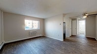 This single-floor unit is thoughtfully laid out to maximize functionality across its key living areas. The main living space features durable wood-style plank flooring, neutral beige walls, and a textured ceiling. A window along the exterior wall allows natural light to filter in while offering outdoor views, and a wall-mounted air conditioning unit alongside baseboard heating ensures year-round comfort.

The galley-style kitchen is efficiently organized with white cabinetry, dark countertops, and black appliances including an electric range, overhead microwave, and double sink. A window at the end of the kitchen brings in additional natural light. The wood-style flooring continues seamlessly from the living area into the kitchen.

The bedroom features soft carpeting, a built-in closet with a sliding door and rod, baseboard heating, and a window that provides natural light and exterior views. An entry area with its own baseboard heater and a white entry door connects the unit to the common hallway.

The bathroom includes a full-size soaking tub with white square tile surround, a small window for ventilation, and standard fixtures. Overall, the unit presents a clean, functional aesthetic suited for everyday living.